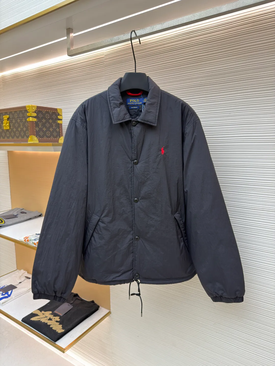 Ralph Lauren red label light cotton jacket (coach jacket) ●Original version: purchased and developed for 2699 ●Item number: 1681910-B90 ●Fabric: high-density nylon taffeta fabric ●Technology: embroidery production ●Color: black ●Version: fitted version ●Weight: about 340 grams ●Size: S-M-L-XL ●Description: The density of high-density nylon taffeta is about 210T-300T, with a smooth and slightly matte surface, excellent windproof and waterproof properties (with DWR water-repellent treatment), strong wrinkle resistance and easy care, in line with the positioning of American casual "durable and practical". Classic dark black, using vat dyeing process (dye penetrates into the fiber), the color fastness reaches ISO level 4 or above, preventing fading or color transfer after washing. The lightweight cotton-polyester blend lining (65% Polyester + 35% Cotton) reduces the friction between the fabric and inner clothing, improving the smoothness of wearing; the blended material combines breathability and durability to avoid the stuffy feeling of pure nylon lining. The classic Coach Jacket has a loose fit with slightly dropped shoulder lines (shoulder width is 1-2cm wider than the standard fit). The length reaches the hips and the sleeves are slightly tapered straight sleeves, taking into account both freedom of movement and casual style. Polo horse logo on the left chest: flat satin stitch embroidery (satin embroidery), embroidery thread: 100% polyester high color fastness embroidery thread (the color number is consistent with the brand's standard red color card), stitch density 12-15 stitches/cm, the outline of the horse logo is clear, and the details of the rider and horse are burr-free. Embroidery positioning: precise positioning through the template (12cm from the neckline and 8cm from the placket) to ensure left and right symmetry and centering. Topstitching technology: The seams of the body are made of double-needle flat seams (needle pitch 3cm/10 needles), the stitches are smooth and straight, the stitching color is dark black (the same color as the fabric), and the stitch width is 0.5cm, which enhances firmness while forming simple decorative lines. The splicing of the front placket and the body: the placket is made of double-layer folding + flat seam splicing, with a thickness of about 0.3cm to ensure that the buttons are not easily deformed when fixed; the edges of the placket are cut with a hot knife to avoid burrs. The sleeves are spliced ​​to the body: the armholes are spliced ​​with round-head keyhole machine seams + flat seams. After splicing, the armholes have a natural curvature and no sense of tightness during movement; the height of the sleeves is about 8-10cm, which is in line with the loose characteristics of casual jackets. Main button: black urea resin button, matte surface treatment, buttonholes are hand-made keyholes (6 strands of cotton thread, keyhole density 8 stitches/mm), and the edges of the eyelets are smooth and without burrs. Collar buttons: The first button on the collar is a hidden button (diameter 12mm), which prevents the collar from opening and improves the neatness of wearing.